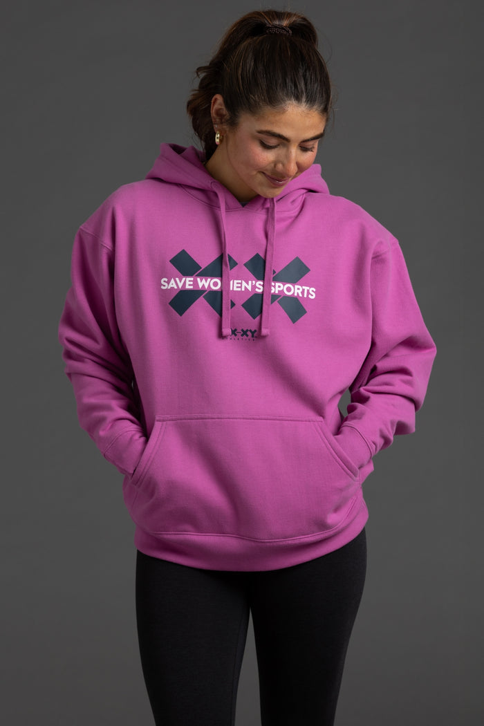 A woman wearing a bright pink pullover hoodie with a dark navy graphic on the chest that reads 'SAVE WOMEN'S SPORTS' and 'SKYX', paired with black leggings, against a solid dark gray studio background. || Bright Fuchsia