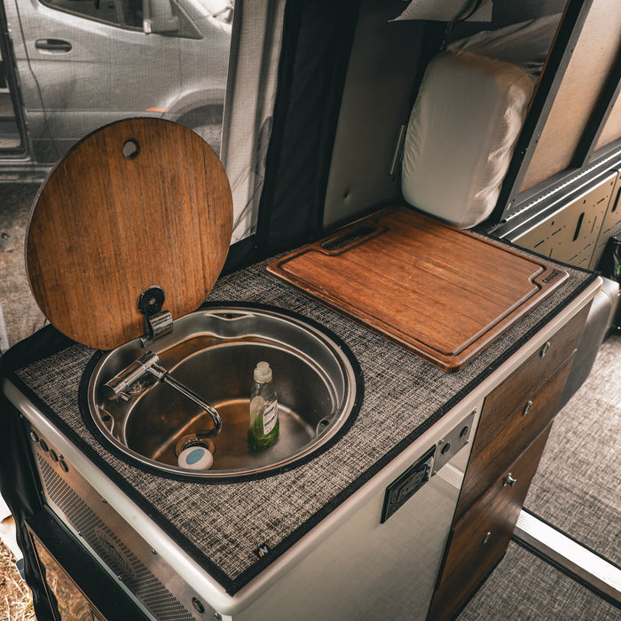 Bamboo sink cover by STO shown inside a van kitchen beside a round metal sink and counter space.