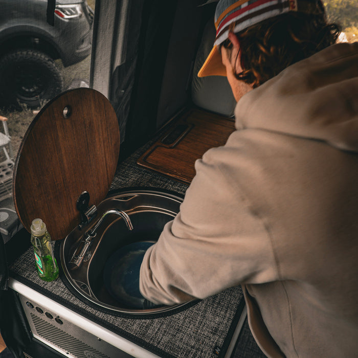 Person washing hands in a round sink with bamboo cover inside a van kitchen setup