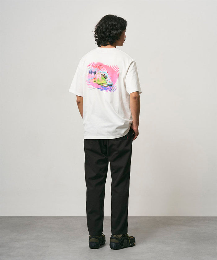 Back view of a model wearing a white Gramicci Bass Tee with a graphic and black pants on a light-tone background.