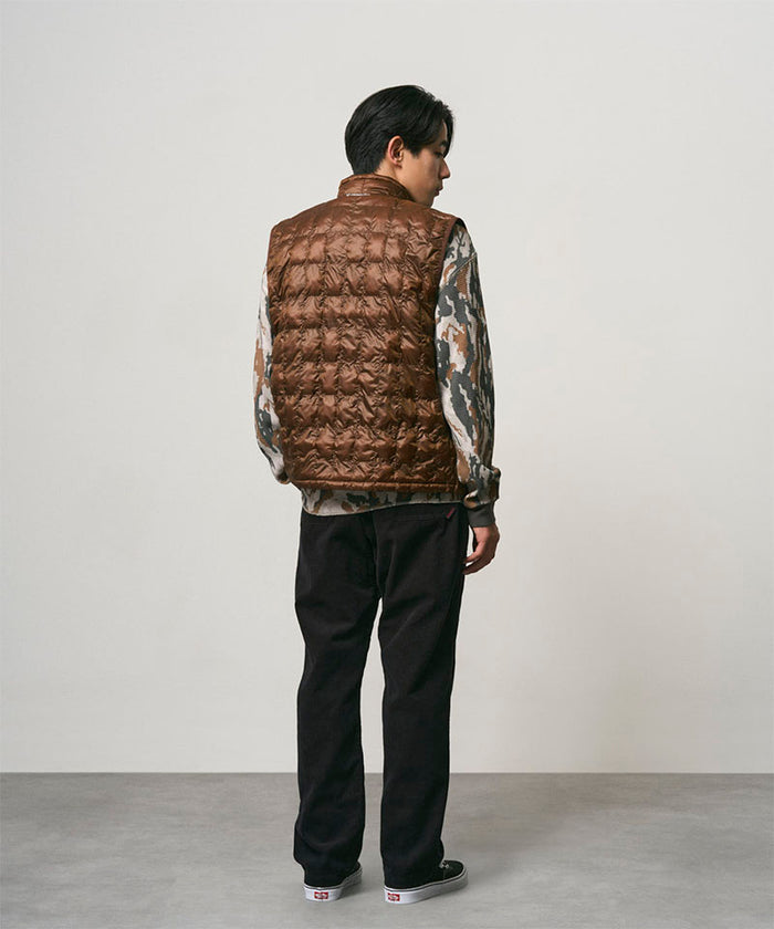 Back view of a model wearing black Gramicci Corduroy Loose Tapered Ridge Pants and a brown vest on a light-tone background.