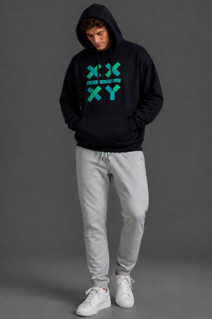 A man wearing a black pullover hoodie with a teal geometric 'XY' graphic on the chest, paired with light gray sweatpants and white sneakers, standing against a dark gray studio background. || Black with Color Logo