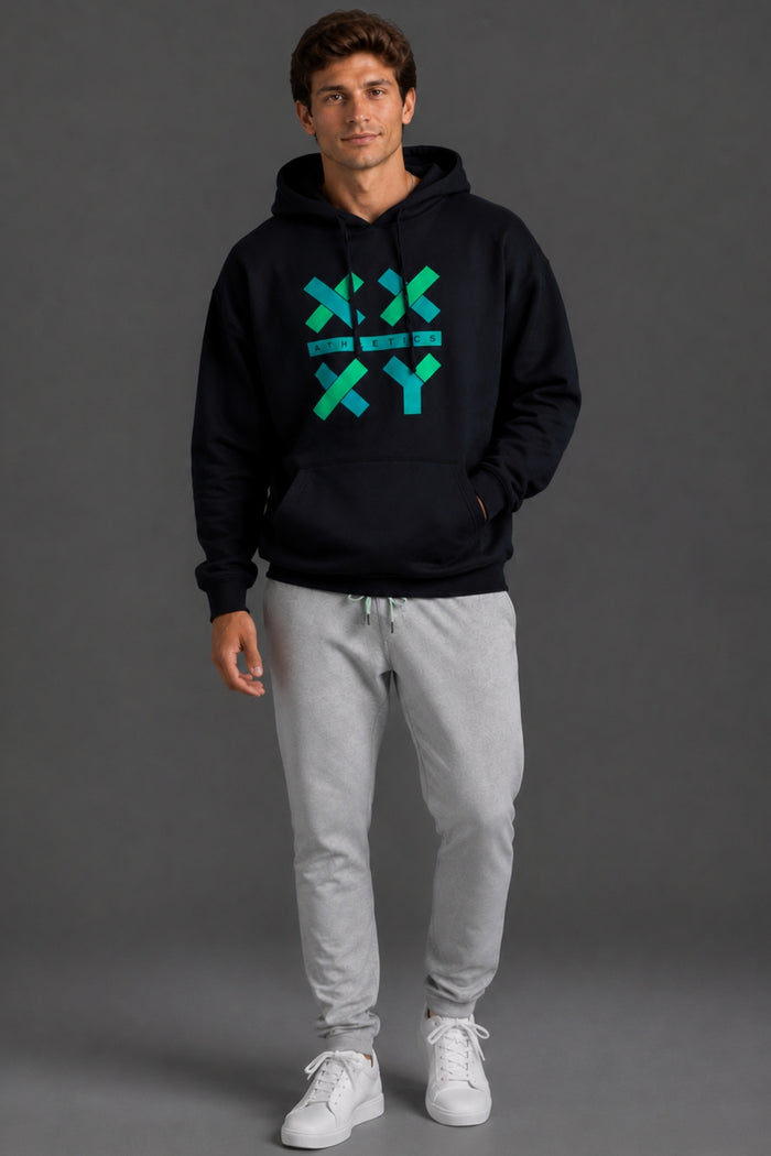 A man modeling a black pullover hoodie with a green and blue geometric 'X' and 'Y' graphic on the chest, paired with light gray sweatpants and white sneakers, against a dark gray studio background. || Black with Color Logo