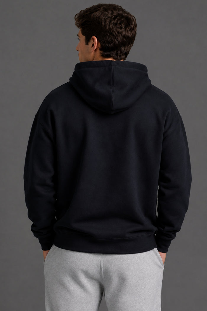 A man viewed from behind wearing a dark navy pullover hoodie and light gray sweatpants against a solid dark gray studio background. || Black with Color Logo