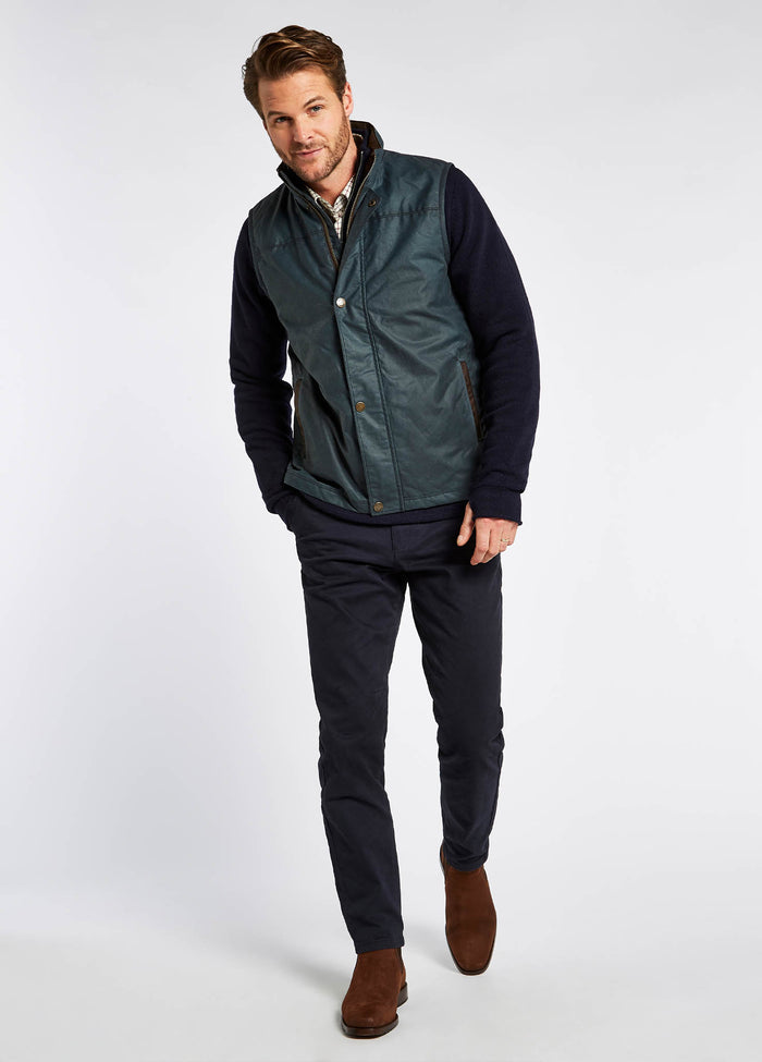 Male model wearing a Dubarry Mayfly wax gilet jacket for men. Dark pebble green blue coloured gilet zipped up with buttons along the zip. The model is also wearing a dark jumper and light shirt underneath the gilet, dark jeans and brown boots.