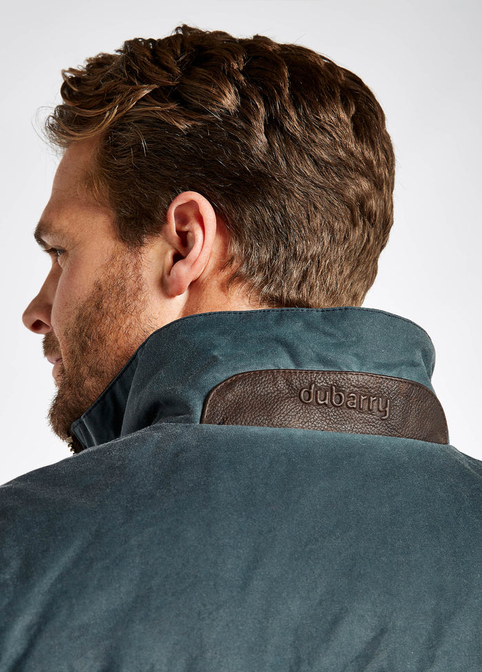 "Close up view of a male model showing the back of a Dubarry Mayfly Wax Jacket for men, dark pebble blue green coloured longer length jacket with a brown leather panel on back of collar. Dubarry is imprinted onto the leather panel of collar
"