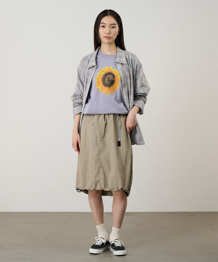 Model wearing a milk tea colored Gramicci Nylon Packable Midi Skirt and plaid shirt over a sunflower tee on a light-tone background.