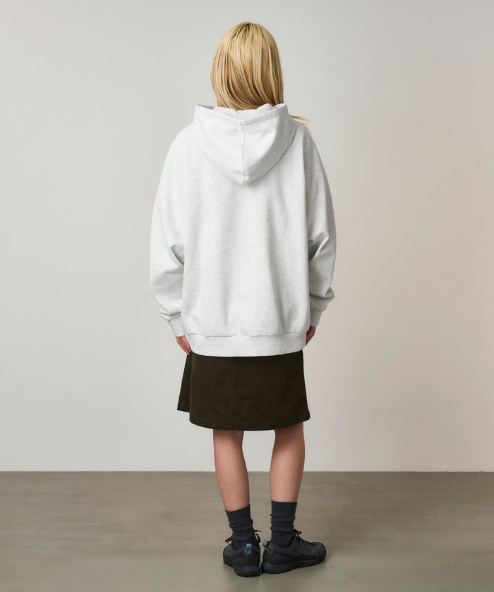 Back view of a model wearing a heather ash colored Gramicci One Point Hooded Sweatshirt and dark skirt on a light-tone background.