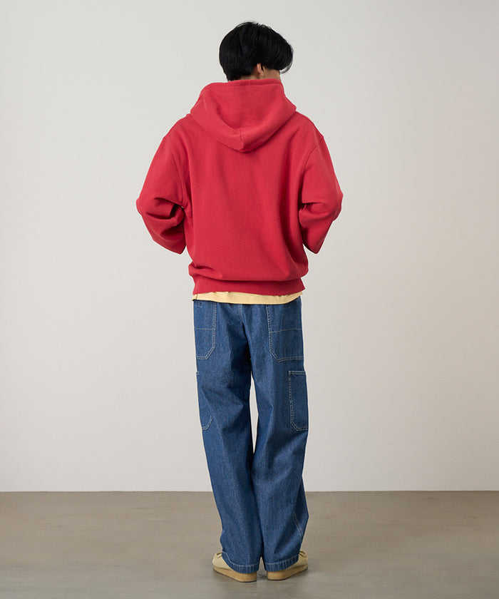 Back view of a model wearing a crimson red Gramicci One Point Zip Hooded Sweatshirt and blue pants on a light-tone background.