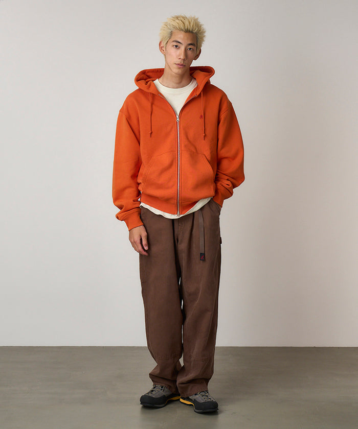 Model wearing a burnt orange Gramicci One Point Zip Hooded Sweatshirt and brown pants on a light-tone background.